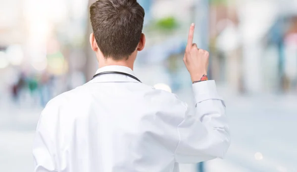 年轻的医生人穿医院外套在孤立的背景摆在后面用手指手指向后面 — 图库照片