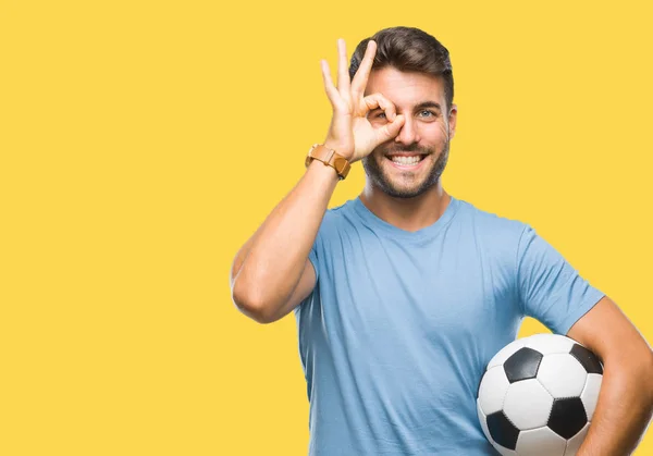 孤立した背景に指を通して見る目に手で サインをして笑っている幸せそうな顔でサッカー サッカー ボールを保持している若いハンサムな男 — ストック写真