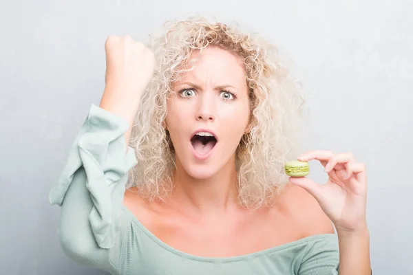 Joven Mujer Rubia Sobre Fondo Gris Grunge Comiendo Macaron Molesto —  Fotos de Stock