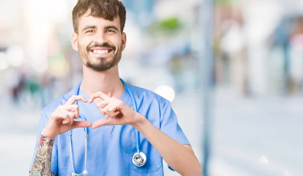 Young handsome nurse man wearing surgeon uniform over isolated background smiling in love showing heart symbol and shape with hands. Romantic concept.