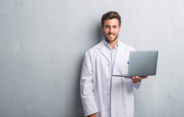 Ayakta ve diş gösterilen kendine güvenen bir gülümsemeyle gülümseyerek mutlu bir yüz ile dizüstü bilgisayar kullanarak üzerinde gri grunge duvar giymiş yakışıklı genç adam beyaz ceket