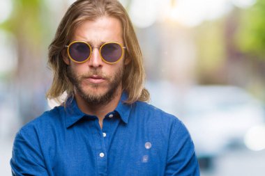 Genç yakışıklı adam uzun saçlı izole arka plan şüpheci ve sinir, onaylamayan ifade yüz çapraz kolları üzerinde güneş gözlüğü takıyor. Negatif kişi.