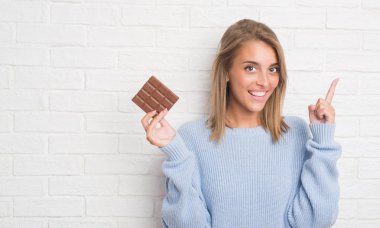 Beyaz tuğla güzel genç kadın çikolatayı bir fikirle sürpriz yemek duvar veya gülen yüz, bir numara ile işaret parmağı soru