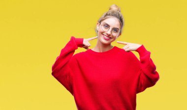 Kendinden emin gösterilen ve işaret parmakları diş ve ağız ile gülümseyen izole arka plan üzerinde kırmızı kazak ve gözlük giyen genç güzel sarışın kadın. Sağlık kavramı.