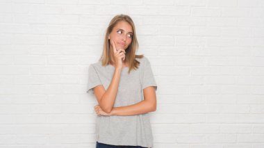 Güzel genç kadın beyaz tuğla duvarın çene düşünme hakkında soru, dalgın ifade üzerine el ile üzerinden. Düşünceli yüzüyle gülümsüyor. Şüphe kavramı.