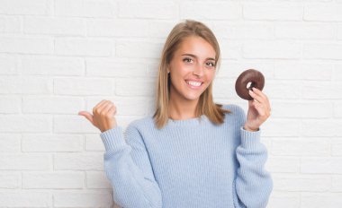 Güzel genç kadın işaret ve başparmak kadar mutlu yüz gülümseyen tarafı ile gösterilen çikolatalı çörek yemek beyaz tuğla duvarın üzerinden