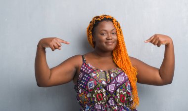 Gri grunge duvar turuncu giymiş genç Afro-Amerikan kadın örgü kendini gururlu ve mutlu parmakları ile işaret emin yüz, gülümseme ile seyir.