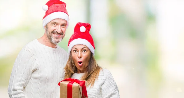 クリスマスの帽子をかぶって 驚き顔 恐怖表現と興奮とショックで怖いの孤立した背景にギフトを保持している中年ヒスパニック系カップル — ストック写真