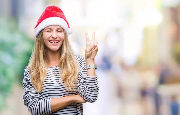 年轻美丽的金发女子戴着圣诞节帽子在孤立的背景微笑与快乐的脸眨眼在相机做胜利标志 第二个 — 图库照片
