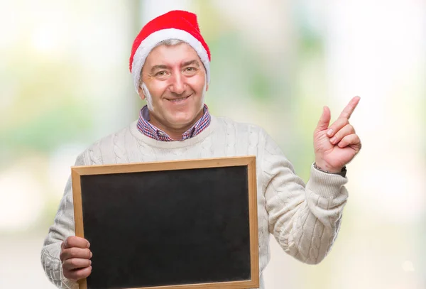 孤立した背景側の手と指で指す非常に幸せにクリスマスの帽子と持株黒板を着てハンサムな年配の男性 — ストック写真