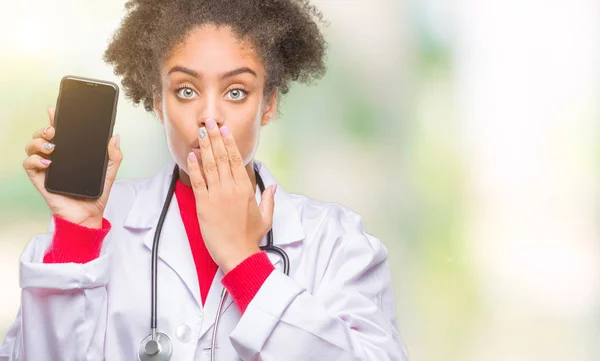 Young afro american doctor woman using smartphone over isolated background cover mouth with hand shocked with shame for mistake, expression of fear, scared in silence, secret concept