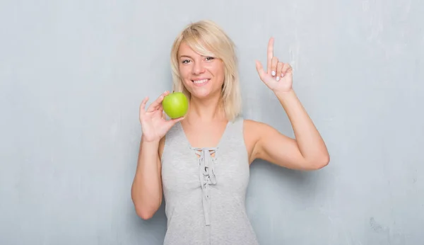 成人白种人妇女在格格灰色墙壁吃绿色苹果惊奇与想法或问题指向手指与愉快的面孔 — 图库照片