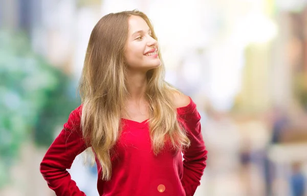 若い美しい金髪女性の自然な表現の顔に笑みを浮かべて側に離れて見て孤立の背景に赤いセーターを着ています 自信を持って笑ってください — ストック写真