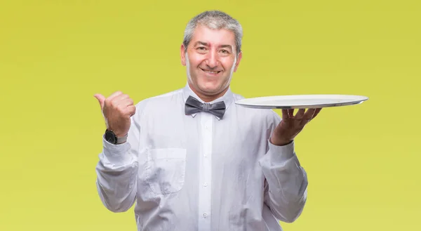 英俊的高级服务员男子拿着银盘在孤立的背景指向和显示与幸福的脸微笑着的一侧 — 图库照片