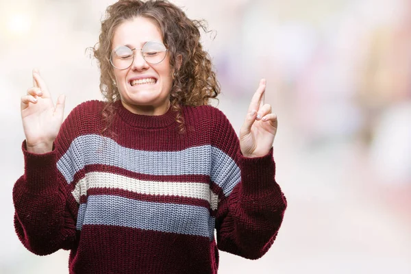 Beautiful brunette curly hair young girl wearing glasses over isolated background smiling crossing fingers with hope and eyes closed. Luck and superstitious concept.