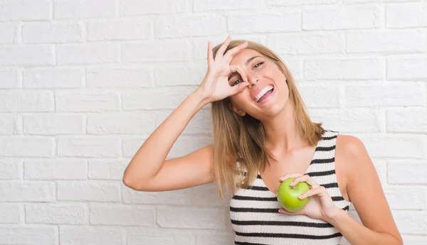 指の間から見て目に手で サインをして笑っている幸せそうな顔で緑を白レンガ壁を食べる以上の美しい若い女性アップル — ストック写真