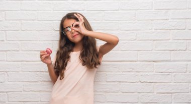 Mutlu yüz el parmaklarının arasından seyir göz üzerinde ok işaretiyle yapıyor gülümseyen ile pembe macaron yeme beyaz tuğla duvarın üzerinden genç İspanyol çocuk