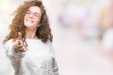 Güzel esmer kıvırcık saçlı Genç kız dostu sunan el sıkışma tebrik ve davet edici olarak gülümseyen izole arka plan üzerinde kış kazak giyiyor. Başarılı iş.