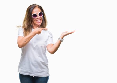 Orta yaş İspanyol kadın hayrete ve el ile sunulması ve parmak ile işaret ederken kameraya gülümseyen izole arka plan üzerinde moda güneş gözlüğü takıyor.