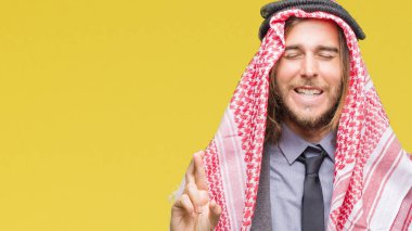 Genç yakışıklı Arap adam uzun saçlı geçiş parmakları umut ve gözleri kapalı ile gülümseyen izole arka plan üzerinde keffiyeh giyiyor. Şans ve batıl kavramı.