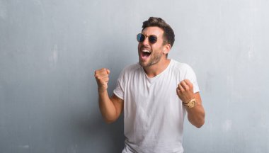 Gülümseyen ve başarı için çığlık yakışıklı genç çok mutlu ve heyecanlı kaldırdı, silah ile kazanan jest yapıyor güneş gözlüğü takıyor gri grunge duvarın üzerinden. Kutlama kavramı.