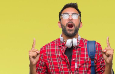 Kulaklıklar ve sırt çantası izole arka plan üzerinde giyen yetişkin İspanyol öğrenci adam hayret ve yukarıya bakıp parmakları ile işaret şaşırttı ve silah kaldırdı.