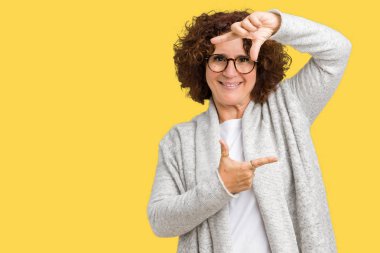 Ceket ve gözlük elle yapma çerçeve ve parmakları ile mutlu yüz gülümseyen izole arka plan üzerinde giyen güzel orta ager üst düzey kadın. Yaratıcılık ve Fotoğrafçılık kavramı.