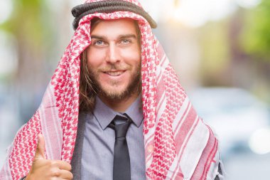 Genç yakışıklı Arap adam uzun saçlı hareketi el ile mutlu yaşasın yapıyor izole arka plan üzerinde keffiyeh giyiyor. Kameraya bakarak başarı gösteren ile ifade onaylama.