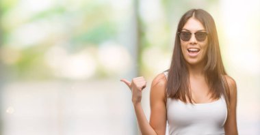 Genç güzel İspanyol seyir ve başparmak yanında yukarıyı mutlu yüz ile gülümseyen güneş gözlüğü takıyor.