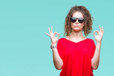 Güzel esmer kıvırcık saçlı Genç kız giyiyor güneş gözlüğü izole arka plan üzerinde dinlenmek ve gülümseyen gözlerle kapalı parmak jestle meditasyon yapıyor. Yoga kavramı.