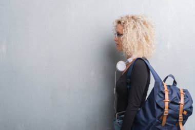 Sarışın genç kadın sırt çantası ve tarafına, seyir kulaklıklar giyen grunge gri duvarın üzerinden dinlenmek doğal yüz emin gülümseme ile pozuyla profili.