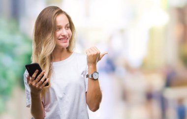 Genç güzel sarışın kadın işaret ve başparmak kadar mutlu yüz gülümseyen tarafı ile gösteren izole arka plan üzerinde akıllı telefon kullanma