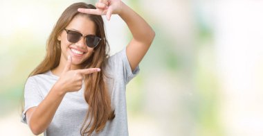 Genç güzel esmer kadın elle yapma çerçeve ve parmakları ile mutlu yüz gülümseyen izole arka plan üzerinde güneş gözlüğü takıyor. Yaratıcılık ve Fotoğrafçılık kavramı.