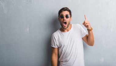 Yakışıklı delikanlı ile başarılı fikir parmak işaret güneş gözlüğü takıyor gri grunge duvarın üzerinden. Çıktıktan ve mutlu. Bir numara.