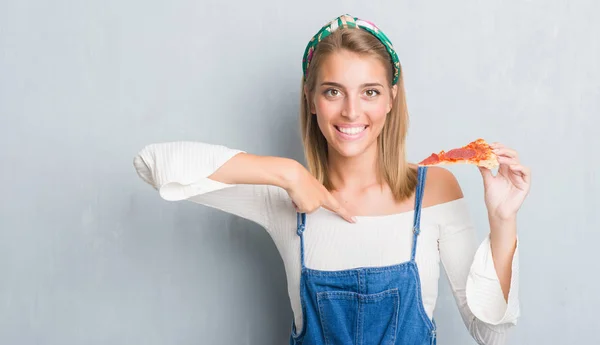 自分自身に驚き顔人差し指とペパロニのピザのスライスを食べるグランジ灰色の壁に美しい若い女性 — ストック写真