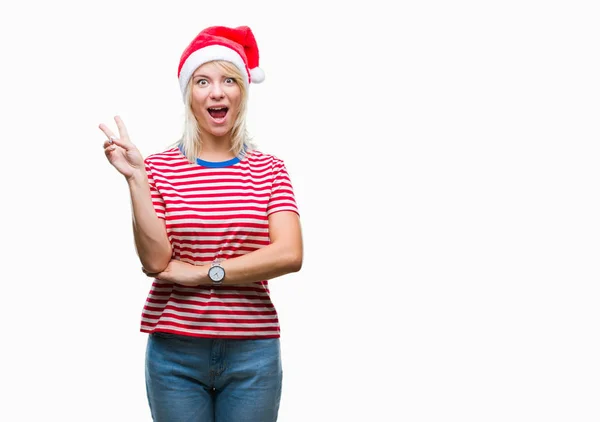 年轻美丽的金发女子戴着圣诞节帽子在孤立的背景微笑与快乐的脸眨眼在相机做胜利标志 第二个 — 图库照片