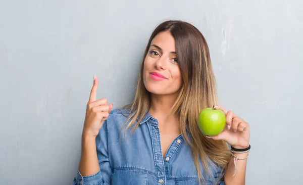 年轻的成年妇女在灰色的垃圾墙吃新鲜绿色苹果惊讶与一个想法或问题指向手指与幸福的脸 — 图库照片
