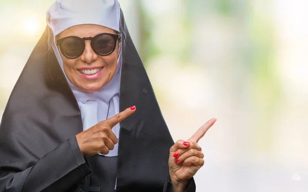 笑顔で側に つの手と指で指しているカメラを見て孤立の背景の上にサングラスをかけている中年シニアのカトリック修道女女 — ストック写真