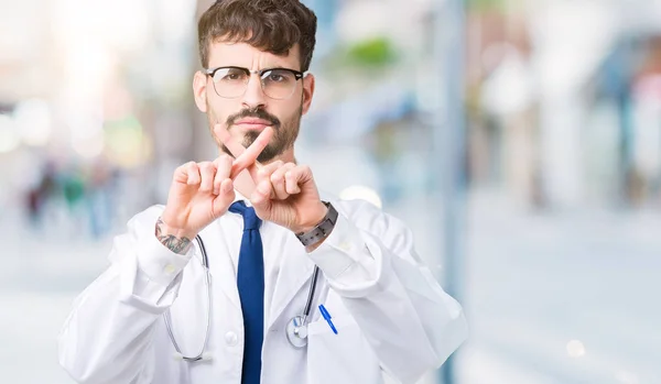 Young doctor man wearing hospital coat over isolated background Rejection expression crossing fingers doing negative sign