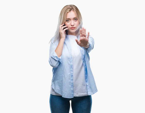 年轻的金发碧眼的女人说话使用智能手机在孤立的背景与开放的手做停止标志与严肃和自信的表情 防御手势 — 图库照片