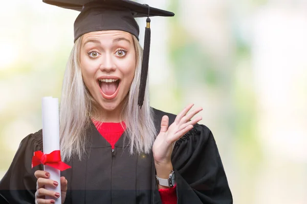 年轻的金发女子穿着研究生制服持有学位在孤立的背景下非常高兴和兴奋 优胜者表达庆祝胜利尖叫着大微笑和举手 — 图库照片