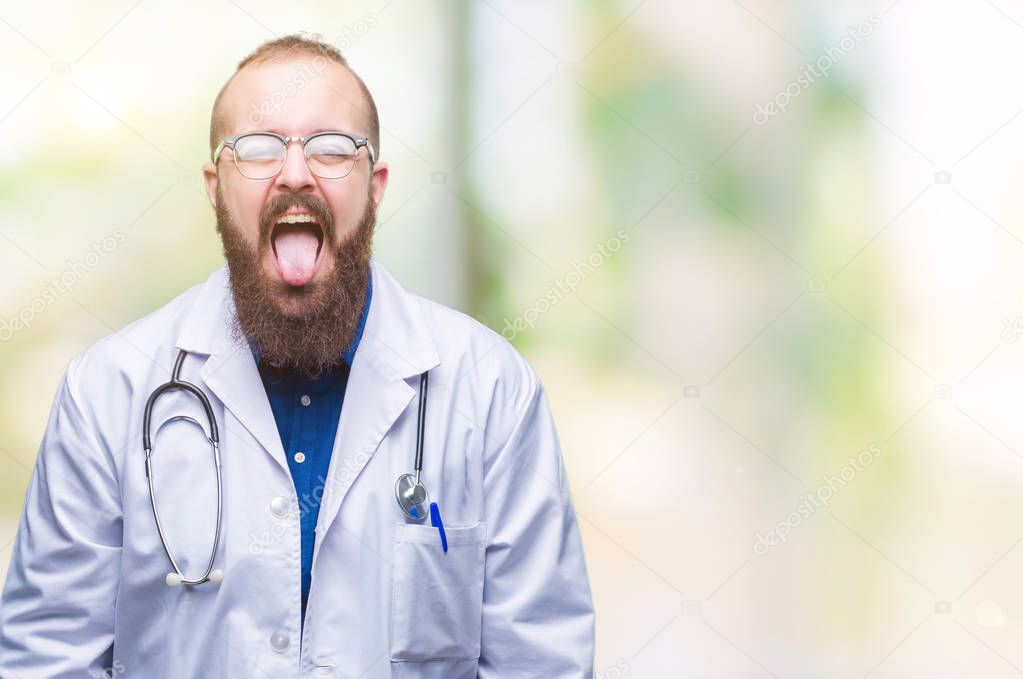 Young caucasian doctor man wearing medical white coat over isolated background sticking tongue out happy with funny expression. Emotion concept.