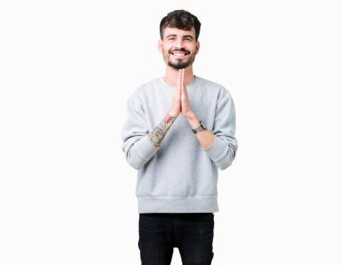 Ellerle birlikte AF emin gülümseyen için soran dua izole arka plan üzerinde sweatshirt giyen genç yakışıklı adam.