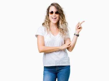 Kameraya bakarak yan el ve parmak ile işaret yüzünde kocaman bir gülümseme ile izole arka plan üzerinde güneş gözlüğü takıyor güzel sarışın genç kadın.