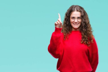 Güzel esmer kıvırcık saçlı Genç kız ve parmak gülümseyen süre bir numara ile kendinden emin ve mutlu yukarıyı gösteren izole arka plan üzerinde gözlük ve kış kazak giyiyor.