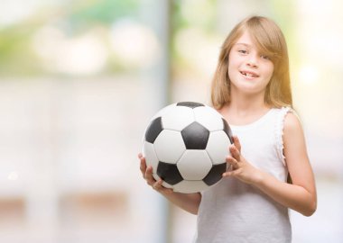 Genç Sarışın yürümeye başlayan çocuk ayakta ve diş gösterilen kendine güvenen bir gülümsemeyle gülümseyerek mutlu bir yüz ile futbol topu tutan