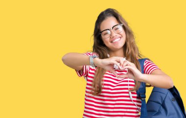 Kalp simgesi ve elleriyle şekil gösterilen aşık gülümseyen izole arka plan üzerinde kulaklık ve sırt çantası giyen genç güzel esmer öğrenci kadın. Romantik kavramı.