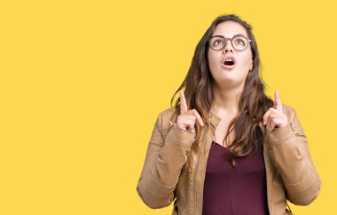 Artı boyutu genç kadın güzel izole arka plan üzerinde elbise ve deri ceket hayret ve yukarıya bakıyor ve parmakları ile işaret ve silah kaldırdı şaşırttı.