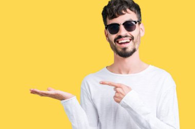 Yakışıklı delikanlı hayrete ve el ile sunulması ve parmak ile işaret ederken kameraya gülümseyen izole arka plan üzerinde güneş gözlüğü takıyor.