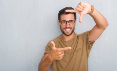 Elle yapma çerçeve ve parmakları ile mutlu yüz gülümseyen gözlüklü gri grunge duvarın üzerinden yakışıklı genç adam. Yaratıcılık ve Fotoğrafçılık kavramı.